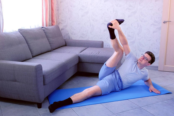 Crazy nerd man is trying to stretch his legs laying on the mat at home. Sport humor concept. - Фото, зображення