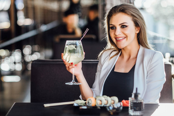 Young, beautiful business woman with lemonsde on hands have a dinner with sushi rolls. - Foto, afbeelding