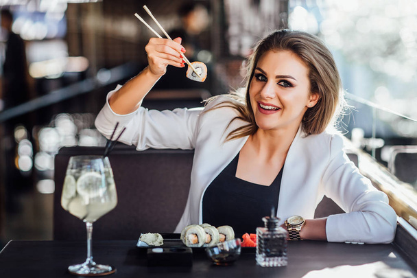 Pretty, young businesswoman eating sushi looking at a fish roll on her chopsticks and posing to the camera. - Foto, afbeelding