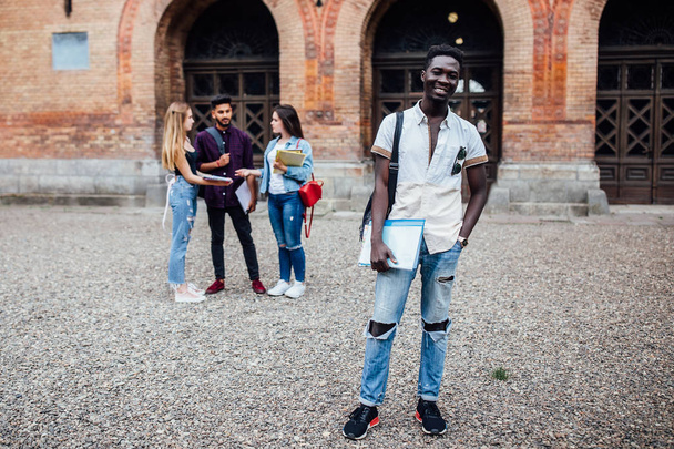 Young, pretty cheerful successful african nerdy student i, standing with books, his classmates on the background, park near campus, sunny day . - Foto, Bild