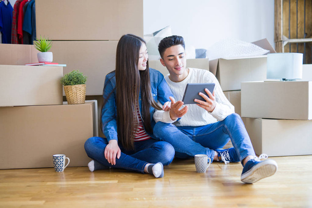 Jovem casal asiático sentado no chão de novo apartamento arround caixas de papelão, usando touchpad tablet e sorrindo em nova casa
 - Foto, Imagem