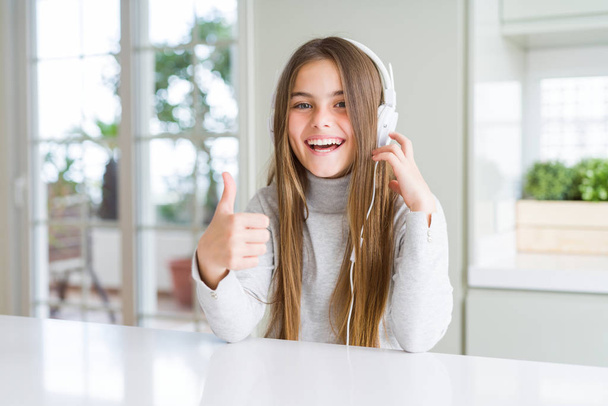 Beautiful young girl wearing headphones listening to music and dancing happy with big smile doing ok sign, thumb up with fingers, excellent sign - Fotografie, Obrázek
