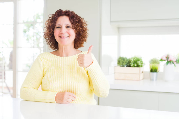 Красива старша жінка в жовтому светрі робить щасливі великі пальці вгору жестом рукою. Затвердження виразу, який показує успіх фотоапарата
. - Фото, зображення