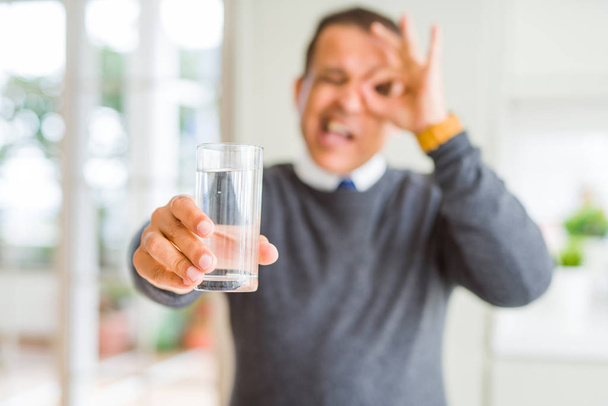 Hombre de mediana edad bebiendo vaso de agua en casa con la cara feliz sonriendo haciendo signo bien con la mano en el ojo mirando a través de los dedos
 - Foto, imagen