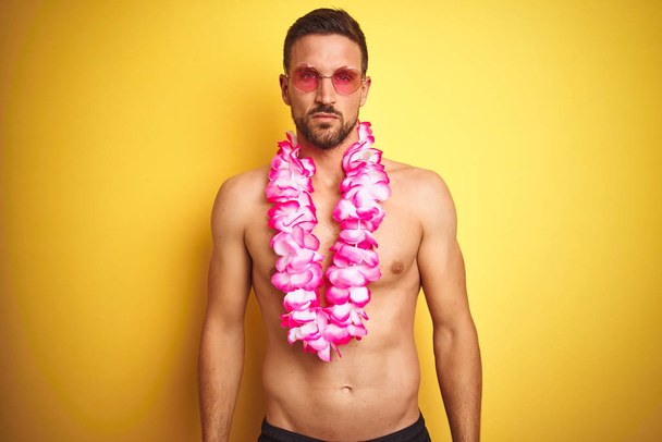 Jeune homme beau torse nu portant des lunettes de soleil et lei hawaïen rose sur fond jaune avec une expression sérieuse sur le visage. Un regard simple et naturel sur la caméra
. - Photo, image