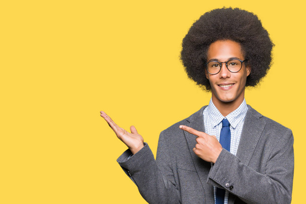 Jeune homme d'affaires afro-américain aux cheveux afro portant des lunettes étonné et souriant à la caméra tout en présentant avec la main et pointant du doigt
. - Photo, image
