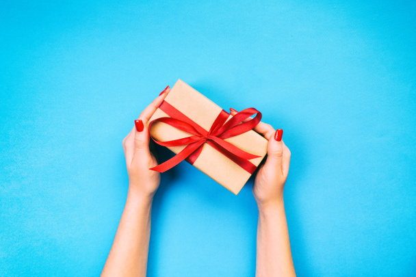 女性の手は、紙に赤いリボンで包まれたバレンタインまたは他の休日の手作りプレゼントを与えます。ボックス、青いテーブルの上のギフト, コピー スペース平面図の装飾 - 写真・画像