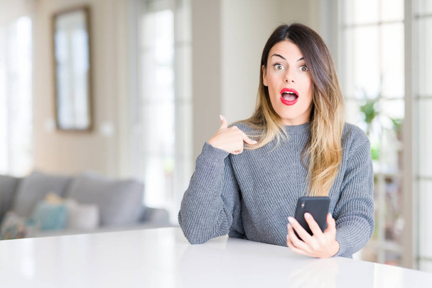 Young beautiful woman using smartphone at home with surprise face pointing finger to himself - Photo, Image