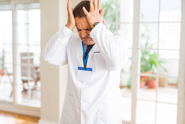 Muž středního věku doktor, který nosí na bílém pozadí lékařský kabát a odznak, který trpí zoufalou bolestí a tlakem, protože bolest a migréna. Ruce na hlavu. - Fotografie, Obrázek