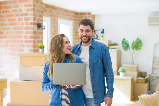 Young couple using computer laptop standing on a room around cardboard boxes, happy for moving to a new apartment - Photo, Image
