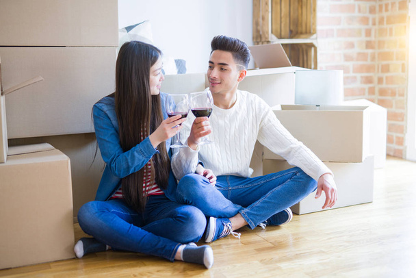 junges asiatisches Paar sitzt auf dem Boden der neuen Wohnung um Pappkartons herum und lächelt und trinkt ein Glas Wein, aufgeregt für den Umzug in ein neues Haus - Foto, Bild