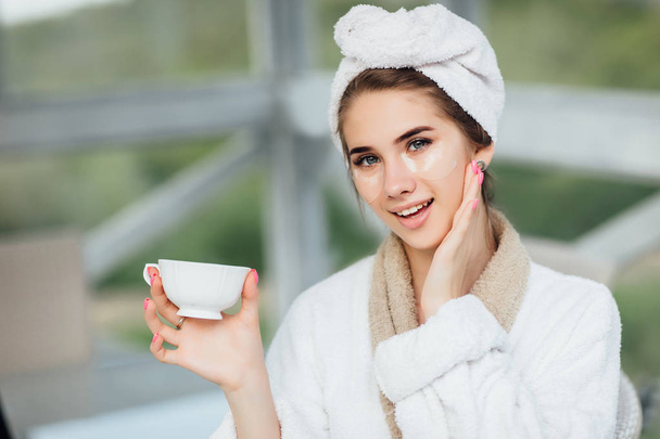 Pretty face. Attractive, smiling girl in white robe, sitting at hotel terrace and holding cup of coffee or tea. Make up concept. - Foto, afbeelding