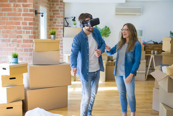 Young couple moving to a new house having fun playing with virtual reality glasses around cardboard boxes - Photo, Image