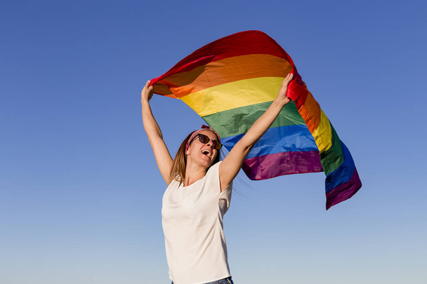 Woman holding the Gay Rainbow Flag over blue sky outdoors. Happiness, freedom and love concept for same sex couples. LIfestyle outdoors - 写真・画像