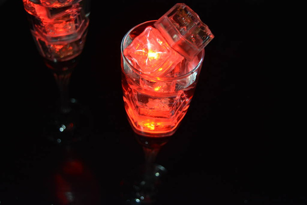 Two unusual glasses of champagne with a drink and bright red ice chips. Plastic ice cubes of ice illuminating the drink in each glass on a black background, an impressive photo and the feast of St. Valentine, the holiday of all lovers. - Fotó, kép