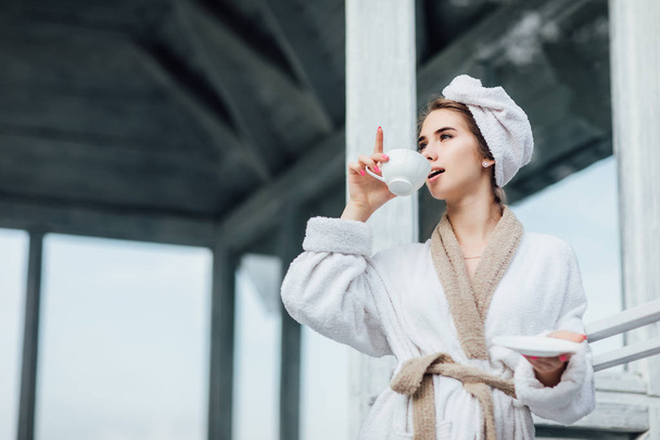 Beautiful,cute young woman wearing at white robe stay on luxury terrace drinking coffee or tea. Vacation in mountains. - Fotografie, Obrázek