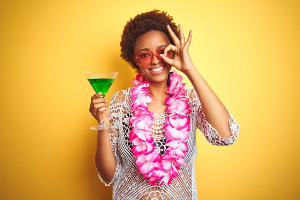 Mujer afroamericana joven con pelo afro vistiendo lei hawaiano flor y bebiendo un cóctel con la cara feliz sonriendo haciendo signo bien con la mano en el ojo mirando a través de los dedos
 - Foto, imagen