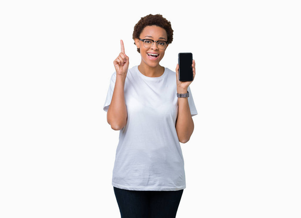 junge afrikanisch-amerikanische Frau zeigt Smartphone-Bildschirm über isoliertem Hintergrund überrascht mit einer Idee oder Frage Zeigefinger mit glücklichem Gesicht, Nummer eins - Foto, Bild