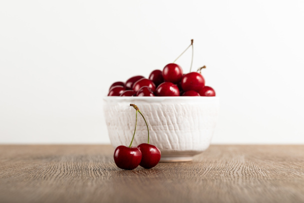 selective focus of red delicious cherries in white bowl and on wooden table isolated on white - Fotografie, Obrázek