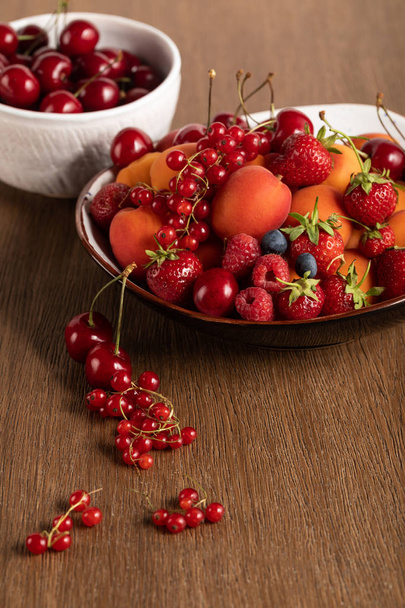 selective focus of red cherries in white bowl and mixed berries on plate on wooden table - Fotografie, Obrázek