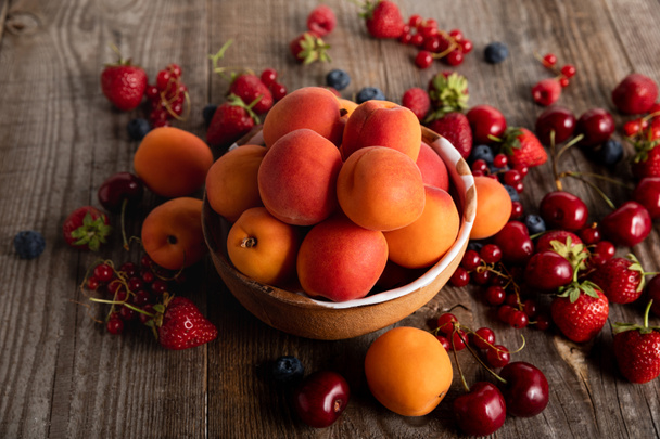ripe seasonal berries scattered around bowl with fresh apricots on wooden table  - Фото, зображення