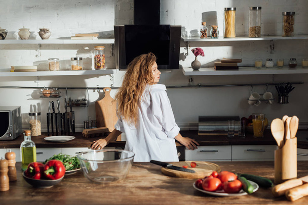 mladá žena v bílé košili, která stála v kuchyni a dívala se jinam - Fotografie, Obrázek