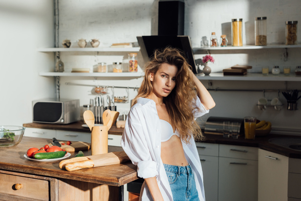 sexy young woman in white bra standing near table and looking at camera in kitchen - Foto, afbeelding