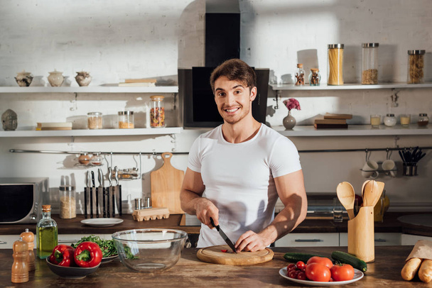mosolyogva izmos férfi, fehér póló vágás zöldségek a konyhában - Fotó, kép