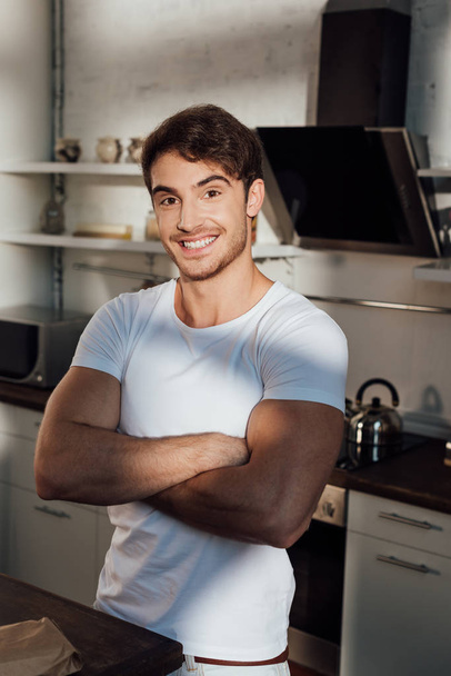 muscular man in white t-shirt standing with crossed arms and smiling in kitchen - Fotó, kép