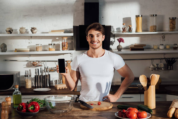 front view of smiling muscular man in white t-shirt showing smartphone with blank screen while cooking in kitchen - Foto, immagini