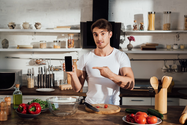 muscular man in white t-shirt pointing with finger at smartphone with blank screen while cooking in kitchen - Foto, immagini