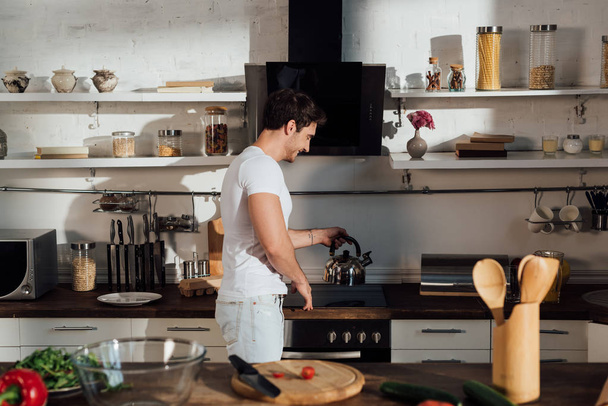 hombre musculoso sonriente en camiseta blanca poner hervidor de agua en el horno en la cocina
 - Foto, Imagen