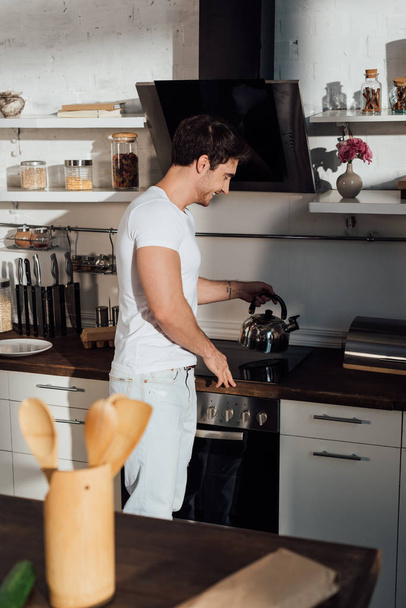 smiling muscular man in white t-shirt putting kettle on oven in kitchen - Φωτογραφία, εικόνα