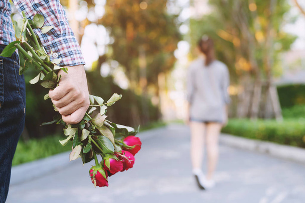 young man standing with a Red Rose on Hand Sadness Love in Ending Break up of Relationship Blurred woman Back Side Walking away parting in public park outdoor. Broken Heart in Valentine day Concept. - Foto, immagini