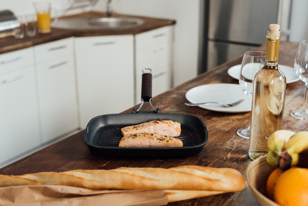 fish in frying pan, fruits, baguettes and wine on table in kitchen - Foto, Imagen