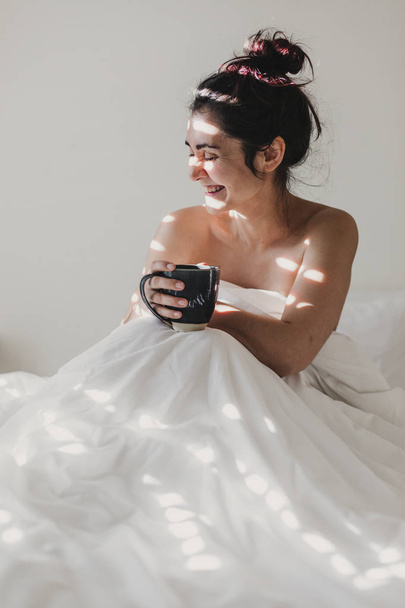 naked woman covered with white sheets sitting on bed and enjoying a cup of coffee. sun is coming through the window. Indoors lifestyle - Zdjęcie, obraz