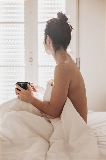 naked woman covered with white sheets sitting on bed and enjoying a cup of coffee. sun is coming through the window. Indoors lifestyle - Foto, Imagen