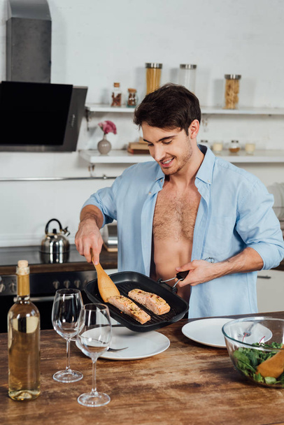 sexy sonriente hombre sosteniendo sartén con pescado en la cocina
 - Foto, imagen