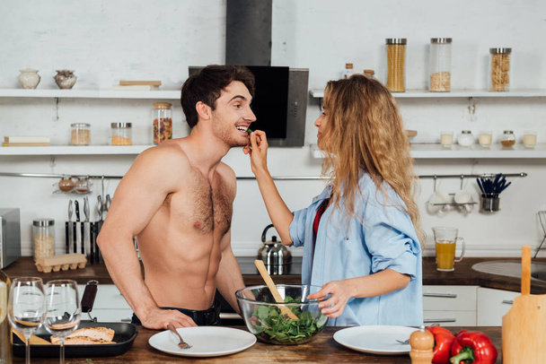 girl feeding shirtless boyfriend with salad in kitchen - Photo, Image