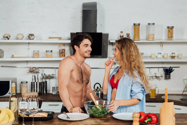 sexy smiling couple standing near table with food and looking at each other in kitchen - 写真・画像