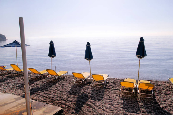 Νωρίς το πρωί στην αμμώδη παραλία χωρίς άτομα με άδεια σεζλόνγκ, ξαπλώστρες, ομπρέλες, καλοκαιρινή ομπρέλα ομπρέλες, ξαπλώστρες, τραπέζι, ξαπλώστρες για ηλιοθεραπεία στην παραλία του ωκεανού περιμένοντας τους τουρίστες. - Φωτογραφία, εικόνα