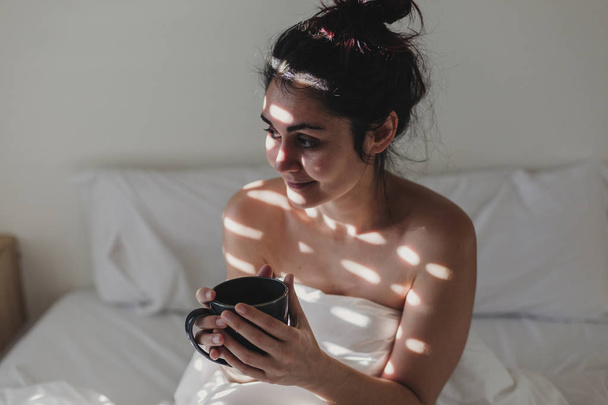 naked woman covered with white sheets sitting on bed and enjoying a cup of coffee. sun is coming through the window. Indoors lifestyle - Photo, Image