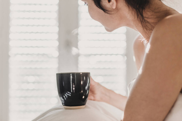 nackte Frau, die mit weißen Laken bedeckt auf dem Bett sitzt und eine Tasse Kaffee genießt. Sonne kommt durch das Fenster. Lebensstil in Innenräumen - Foto, Bild