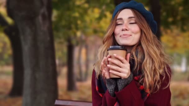 Close up view of Smiling brunette woman in beret hat and coat drinking coffee while sitting on bench in park - Video
