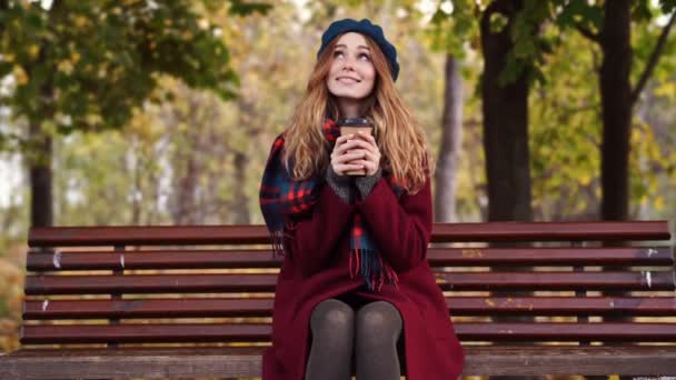 Smiling brunette woman in beret hat and coat drinking coffee while sitting on bench in park - Кадри, відео