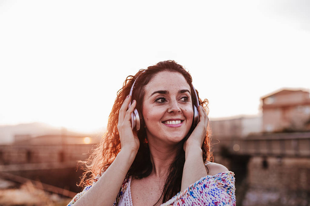 portrait outdoors of a young beautiful woman at sunset listening music on headset and smiling. Lifestyle and music concept - Foto, imagen