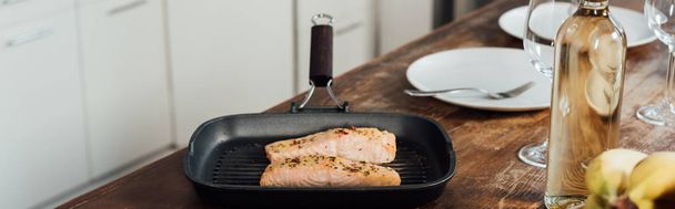 plan panoramique de poêle à frire avec du poisson, bouteille de vin, verres à vin et assiettes sur la table dans la cuisine
 - Photo, image