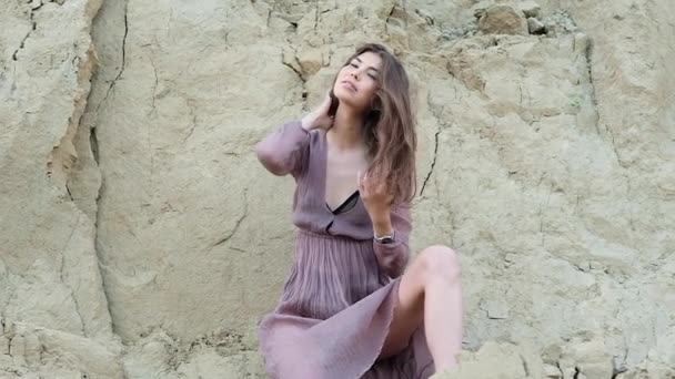 young beautiful slender brunette in a transparent brown dress, black retro swimsuit and dark shoes posing against a sandy cliff. Ruffles her hair and leans forward, looking at the camera - Footage, Video