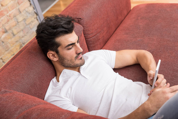 A serious young man laying on a sofa and using his smartphone - Foto, Imagem