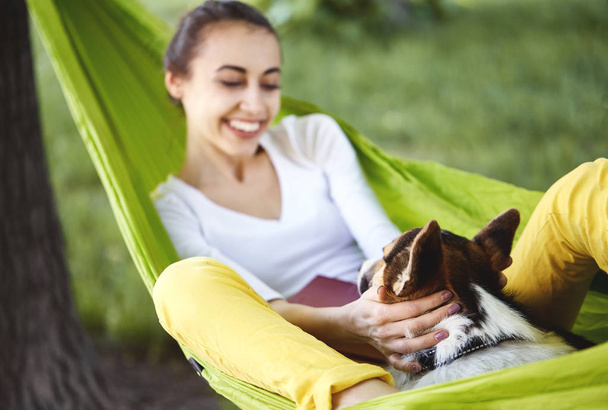 Smiling young woman in green hammock with cute dog Welsh Corgi in a park outdoors. Beautiful happy female in white shirt enjoying good day and hugging a pet. Focus on the dog. - Photo, image